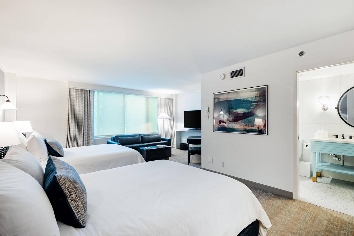 A hotel room with two beds, a desk, a TV, a painting, and a bathroom.