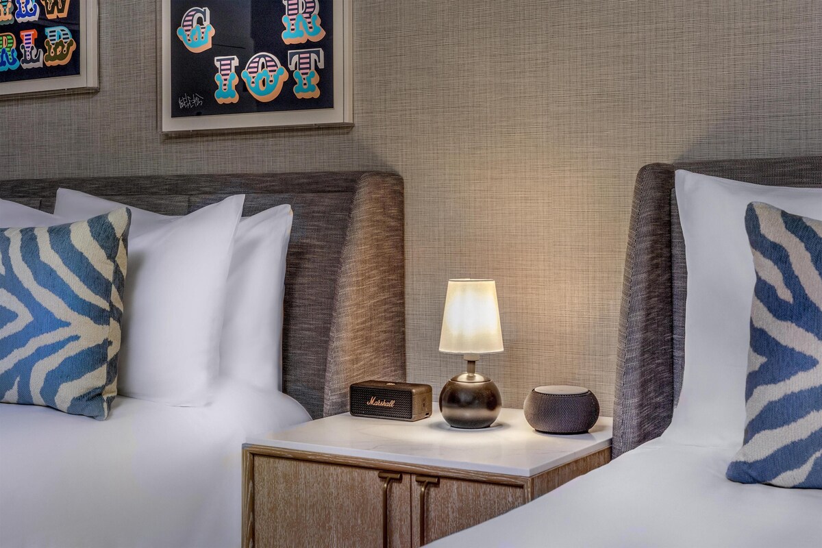 A hotel room with a bed, a nightstand with a lamp, a small speaker, and a framed artwork on the wall.
