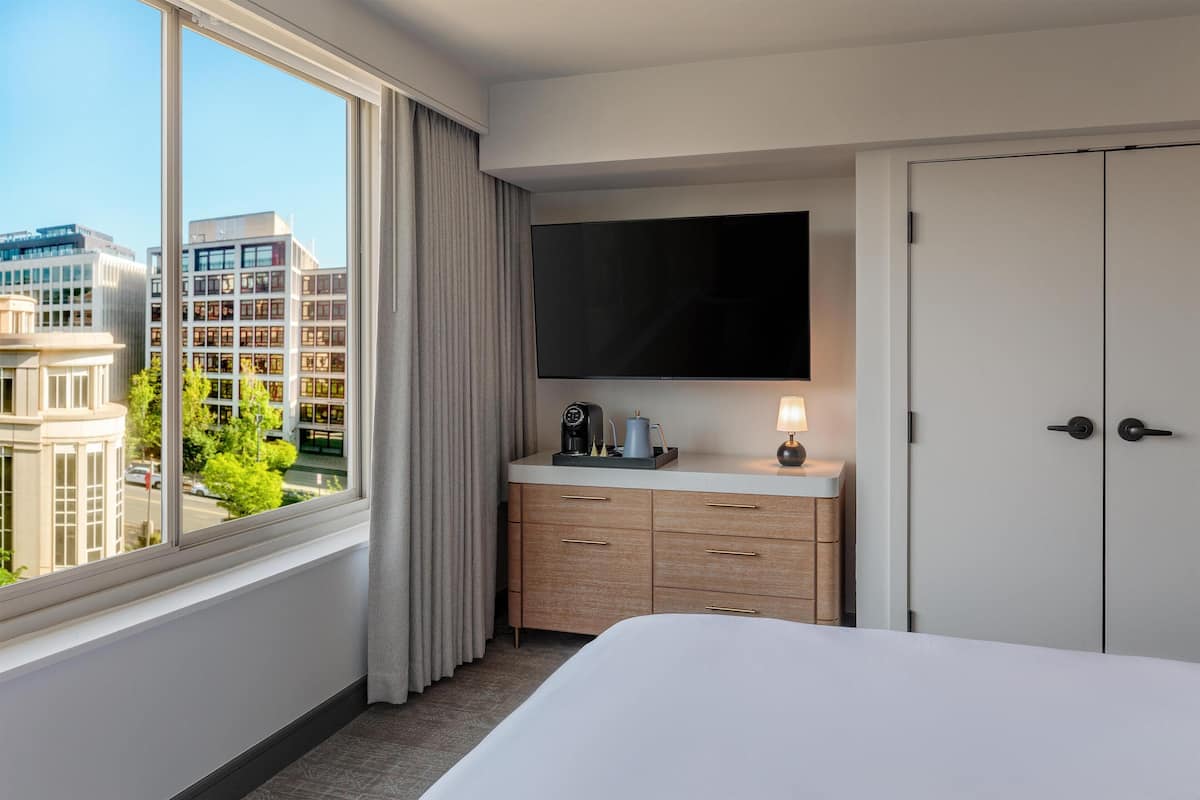 A hotel room with a large window, a bed, a TV, a desk with drawers, and a wardrobe.