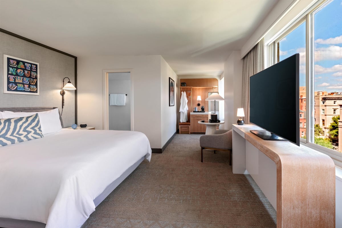 A hotel room with a large bed, a desk with a flat-screen TV, a chair, and a view of the city through large windows.