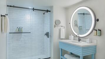 A bathroom with a glass shower enclosure, a white sink with a blue vanity, and a round mirror with a light.