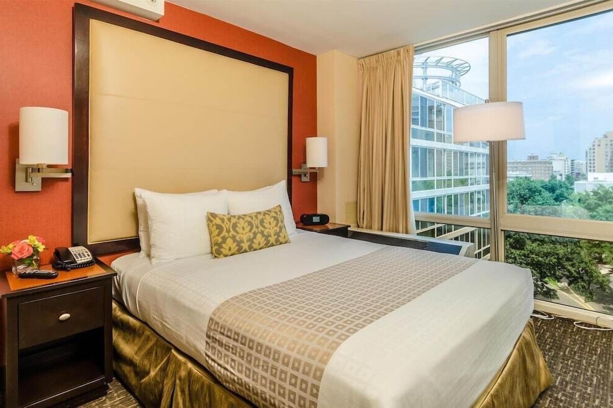 A hotel room with a large bed, a nightstand with a phone and lamp, and a view of a cityscape through a large window.