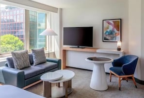 A modern hotel room with a sofa, two side tables, a flat-screen TV, and a large window offering a city view. - The Beacon at Embassy Row Washington DC, Tapestry by Hilton (Washington)