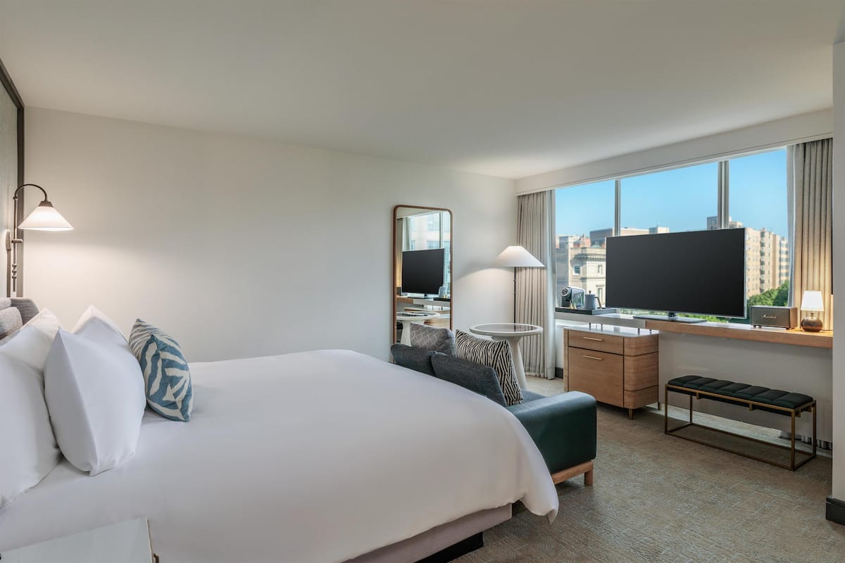 A hotel room with a bed, a desk with a computer, a TV, and a window with a city view.