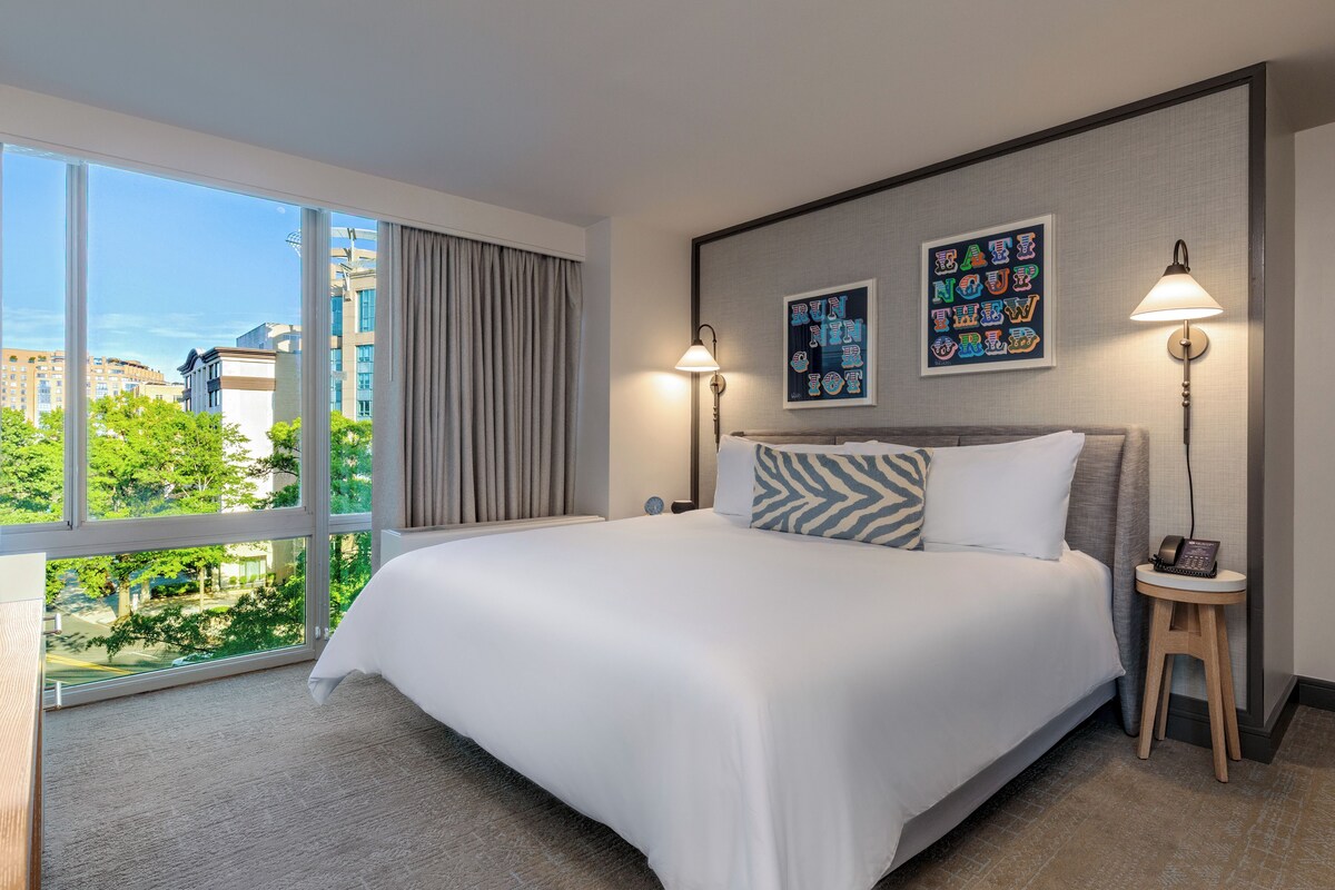 A hotel room with a large bed, a bedside table with a lamp, a view of the city, and a framed picture on the wall.