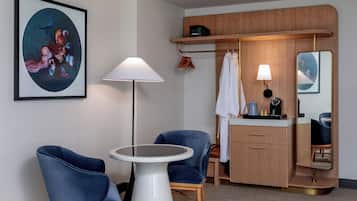A hotel room with a round white table, two blue chairs, a wooden cabinet, a mirror, and a framed abstract painting on the wall.