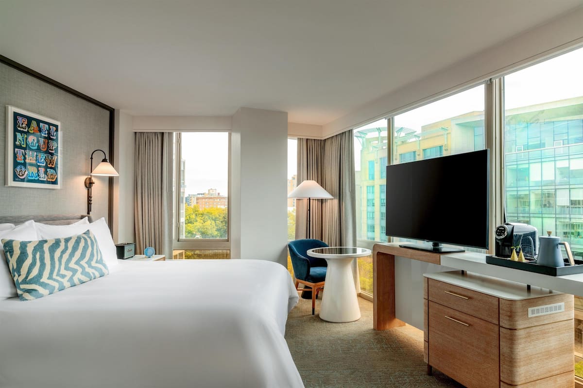 A hotel room with a bed, a desk with a flat-screen TV, a chair, and a view of buildings through large windows.