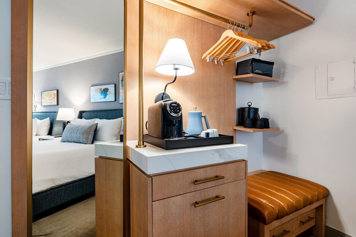 A compact hotel room with a bed, a desk with a lamp, a coffee maker, and a small seating area.