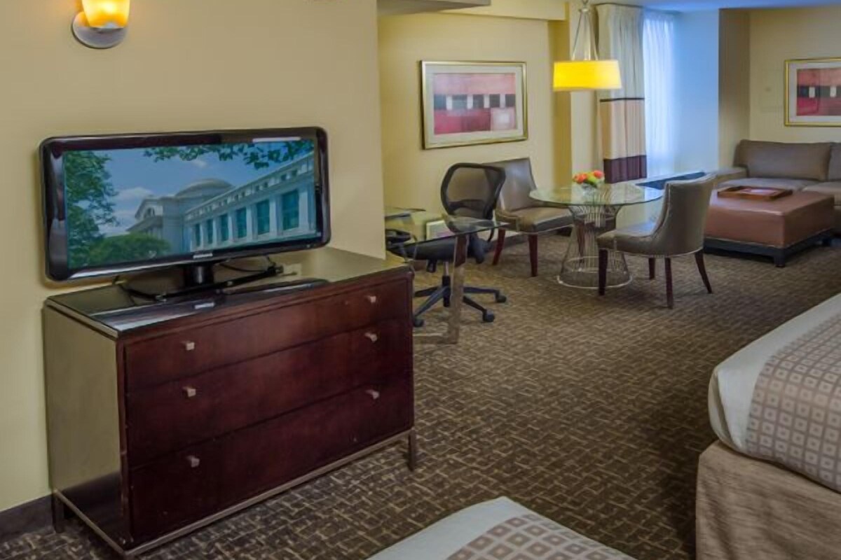 A hotel room with a television on a dresser, a desk with chairs, a sofa, and a patterned rug.