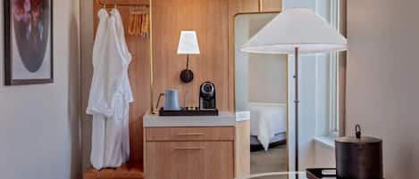 Habitación de hotel compacta con un armario de madera, una mesita de noche con lámpara, una mesa redonda con lámpara y una cama con edredón blanco.