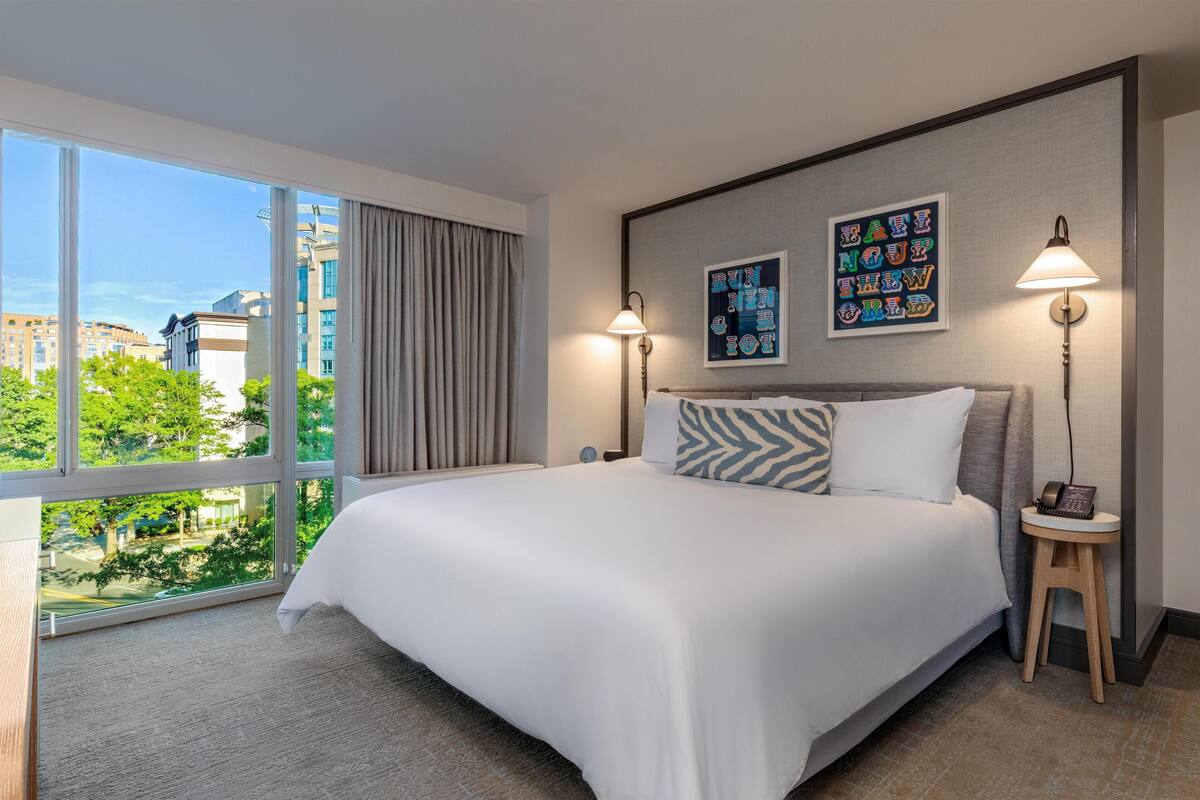 A hotel room with a large bed, two bedside lamps, a view of the city, and a framed artwork on the wall.