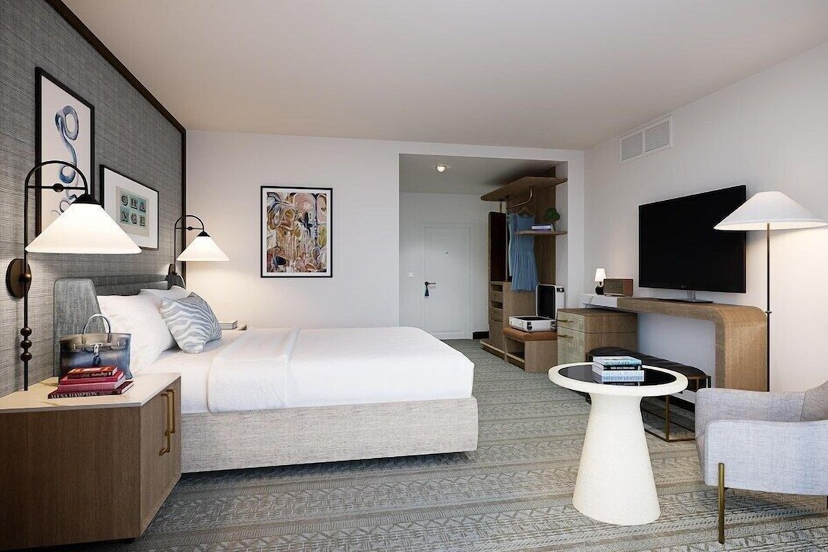 A hotel room with a bed, bedside tables, a TV, a chair, and a small table.