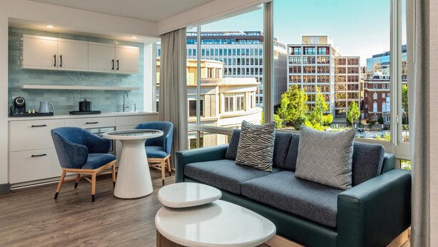 A modern living room with a sofa, two side tables, and a kitchen area with cabinets and a window overlooking a cityscape. - The Beacon at Embassy Row, WorldHotels Distinctive (Washington)