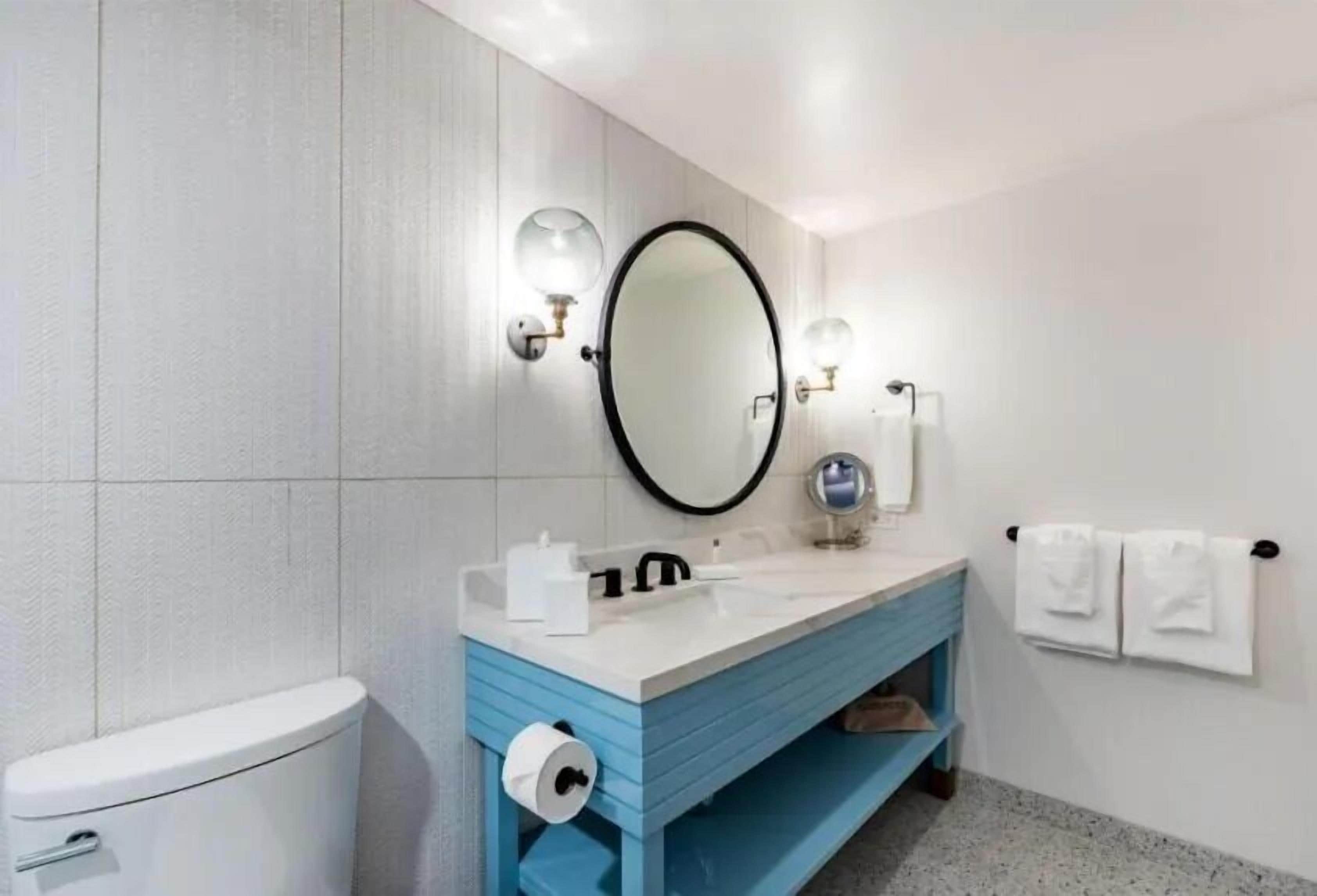 Un baño moderno con inodoro blanco, tocador azul, espejo redondo y un perchero para toallas.