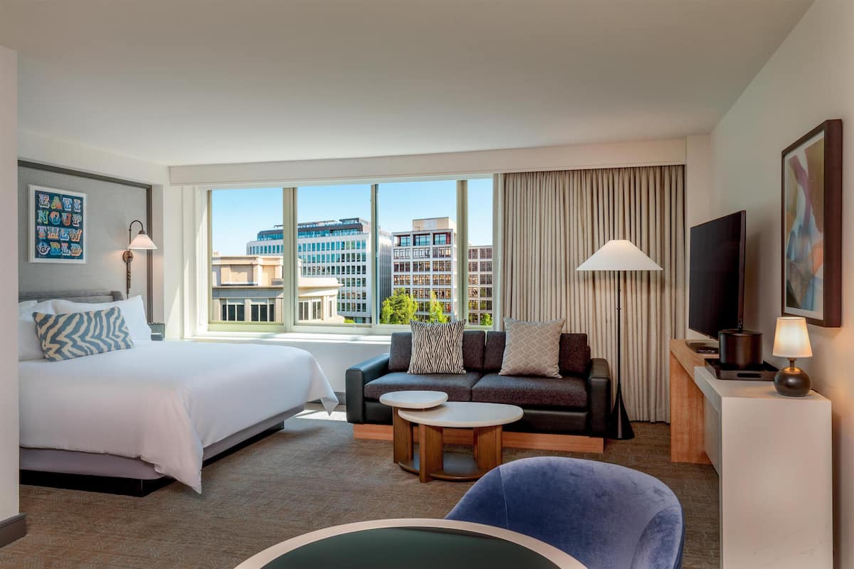 A hotel room with a bed, sofa, two small tables, a TV, and a large window with a city view.