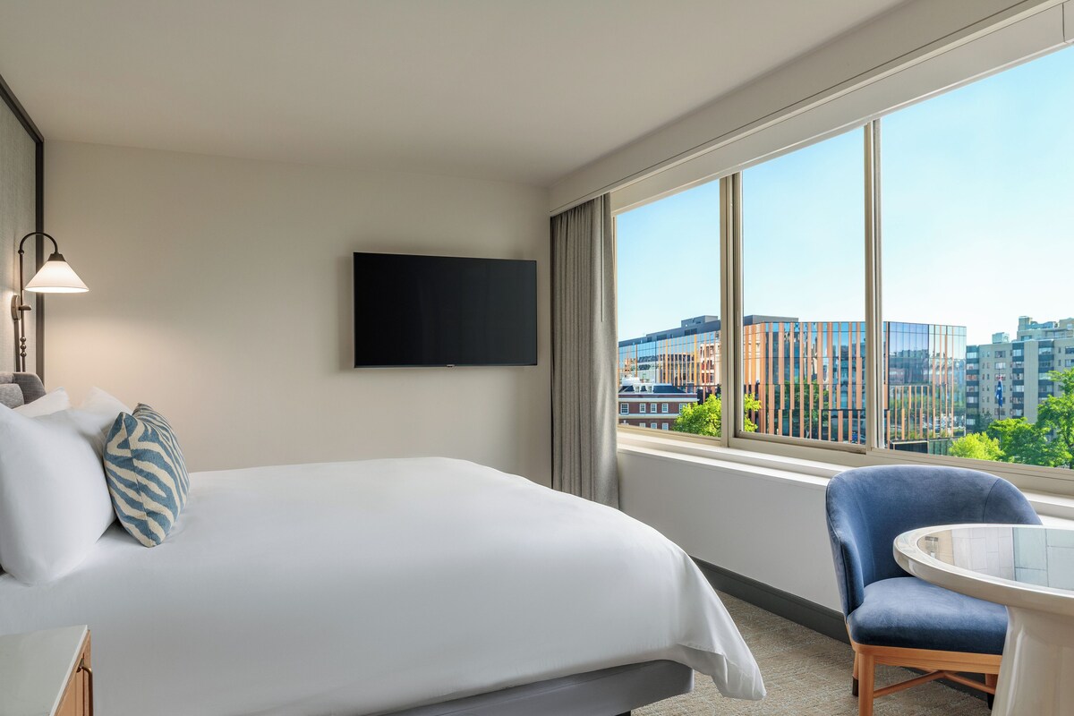 A hotel room with a bed, a TV, a chair, and a window with a city view.