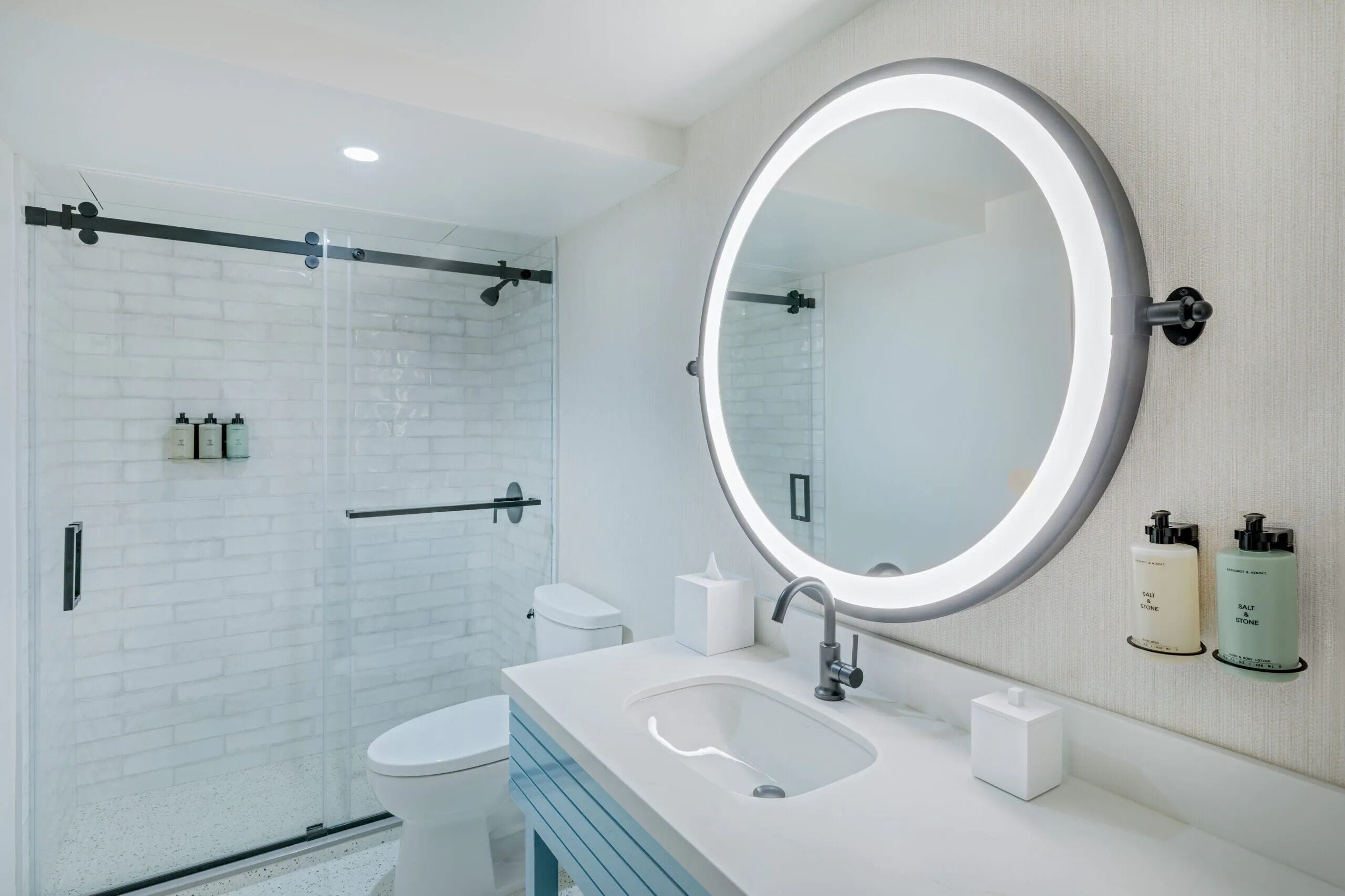 Une salle de bain moderne avec une douche à parois de verre, des toilettes blanches, un lavabo avec un miroir et deux distributeurs de savon.