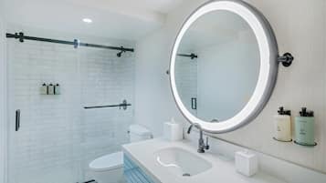 Une salle de bain moderne avec une douche à parois de verre, des toilettes blanches, un lavabo avec un miroir et deux distributeurs de savon.