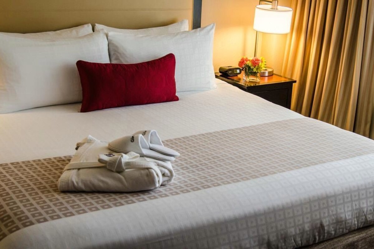 A neatly made bed with white and red pillows, a folded towel, and a bedside table with a lamp and a small vase with flowers.