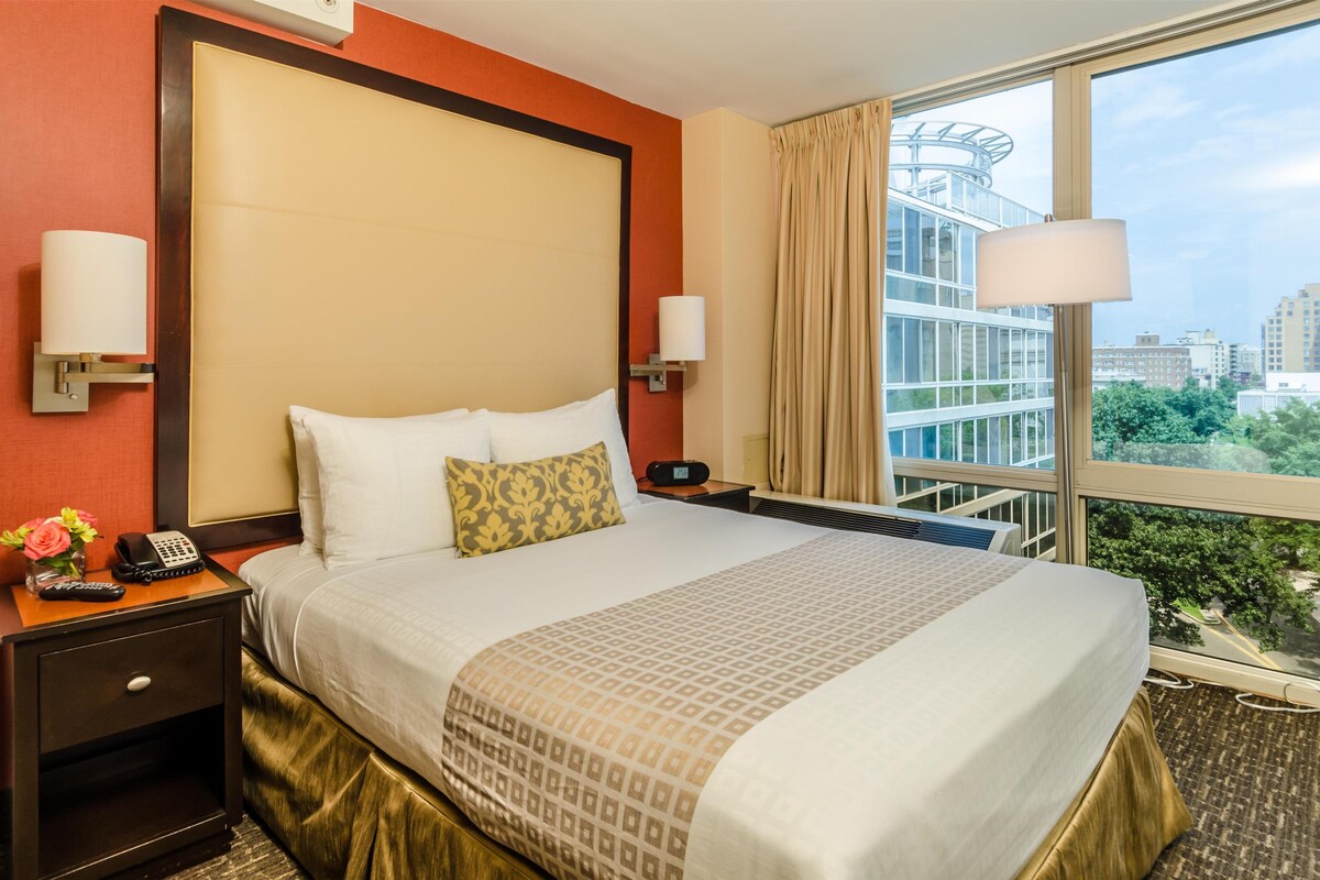A hotel room with a large bed, a nightstand with a phone and remote, a window with a view of buildings and trees, and a lamp on the nightstand.