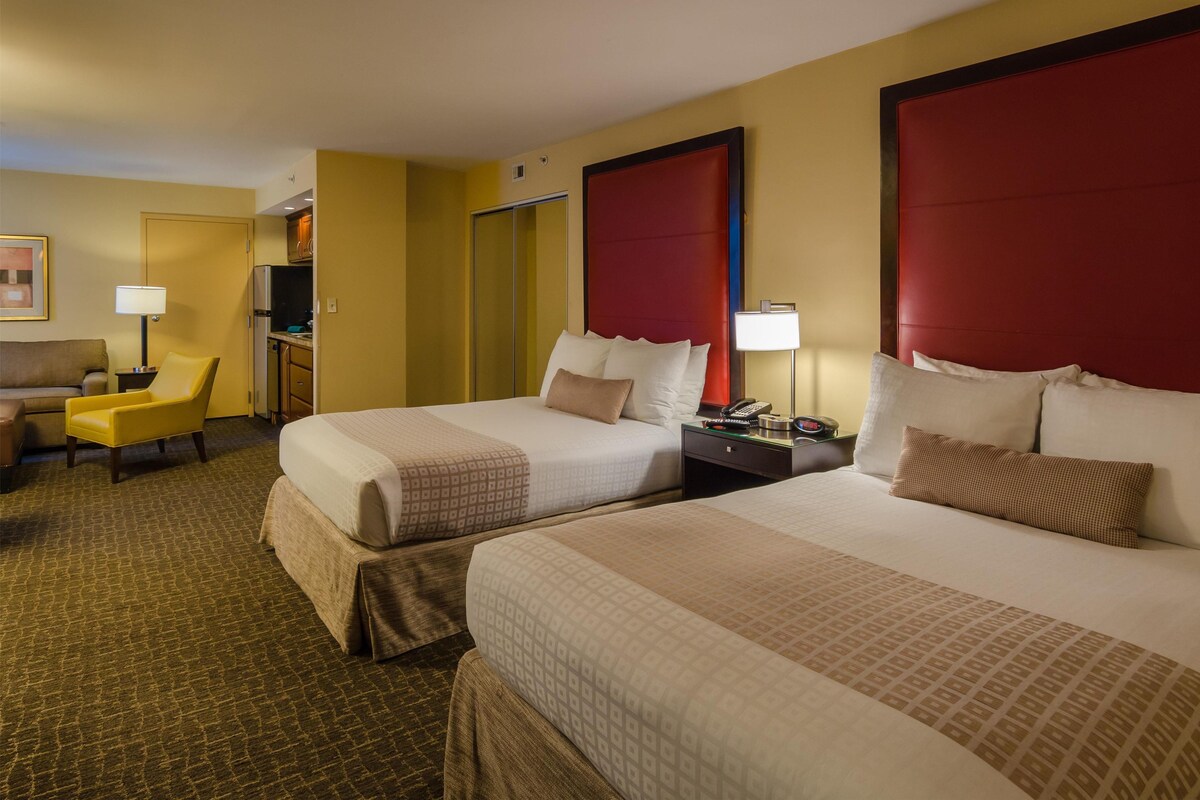 A hotel room with two beds, a desk, a chair, and a small kitchenette.