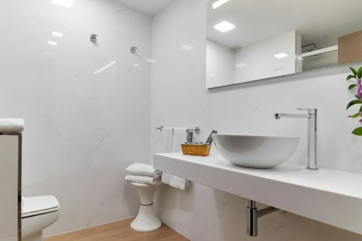 Un baño moderno con inodoro blanco, un lavabo blanco de gran tamaño, un espejo sobre el lavabo y una planta pequeña a la derecha.