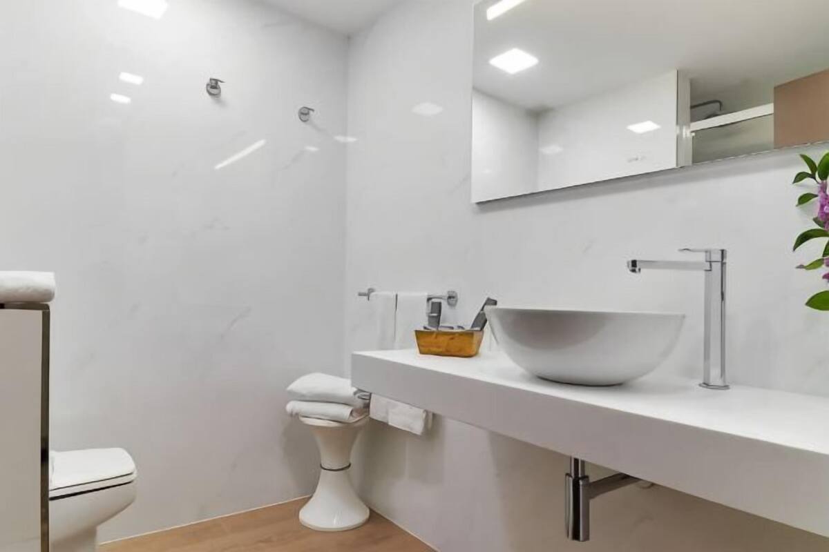 Een moderne badkamer met een wit toilet, een witte wastafel met een grote kom, een spiegel boven de wastafel en een kleine plant aan de rechterkant.