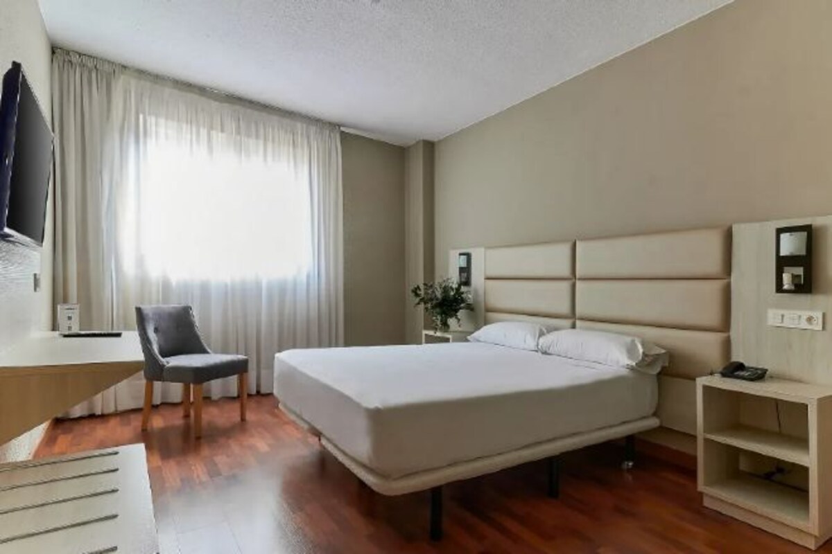 Habitación de hotel con cama, escritorio, silla, mesita de noche, televisor y ventana con cortinas.