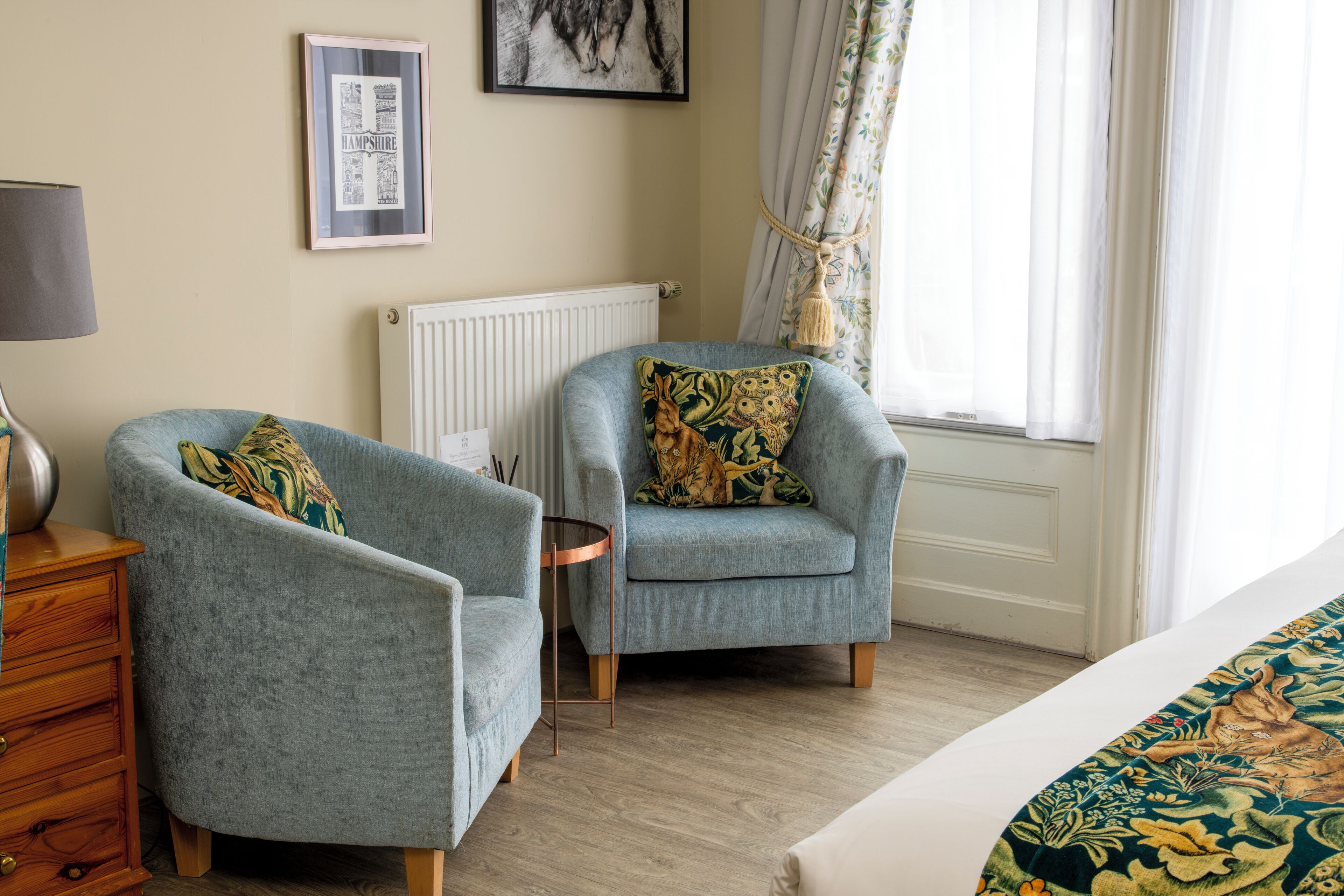 Une pièce comprenant deux fauteuils, une commode en bois, un radiateur et une fenêtre avec des rideaux.