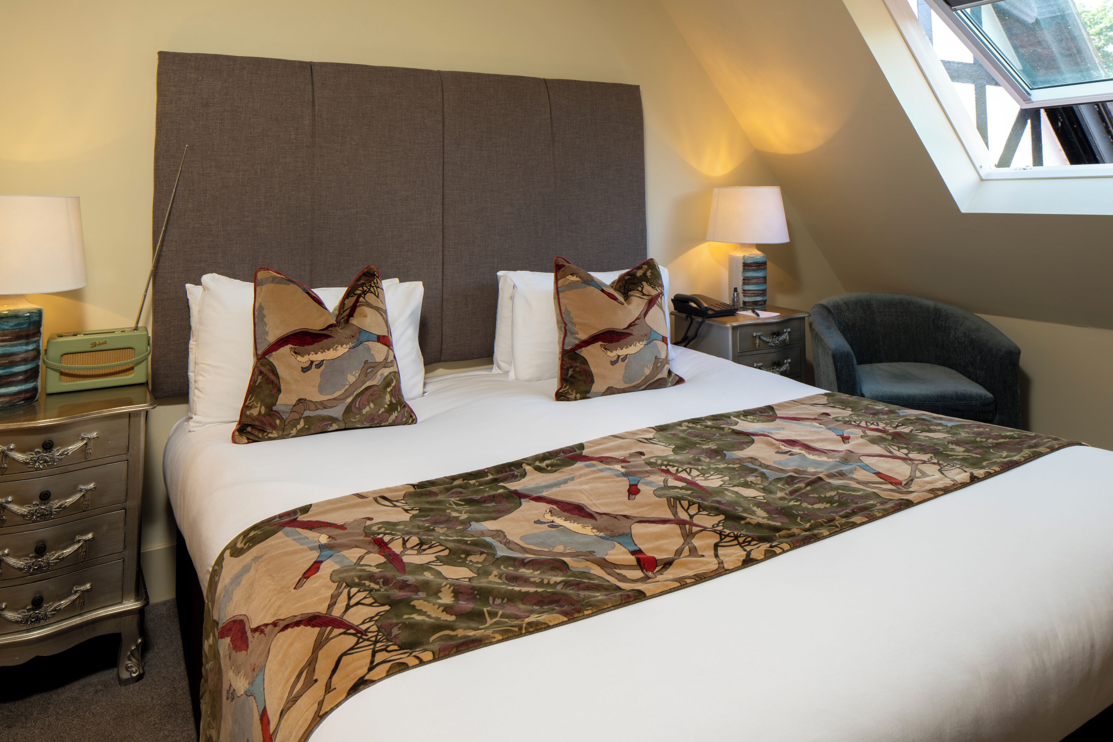 A hotel room with a large bed, two decorative pillows, a nightstand with a lamp, a chair, and a skylight.