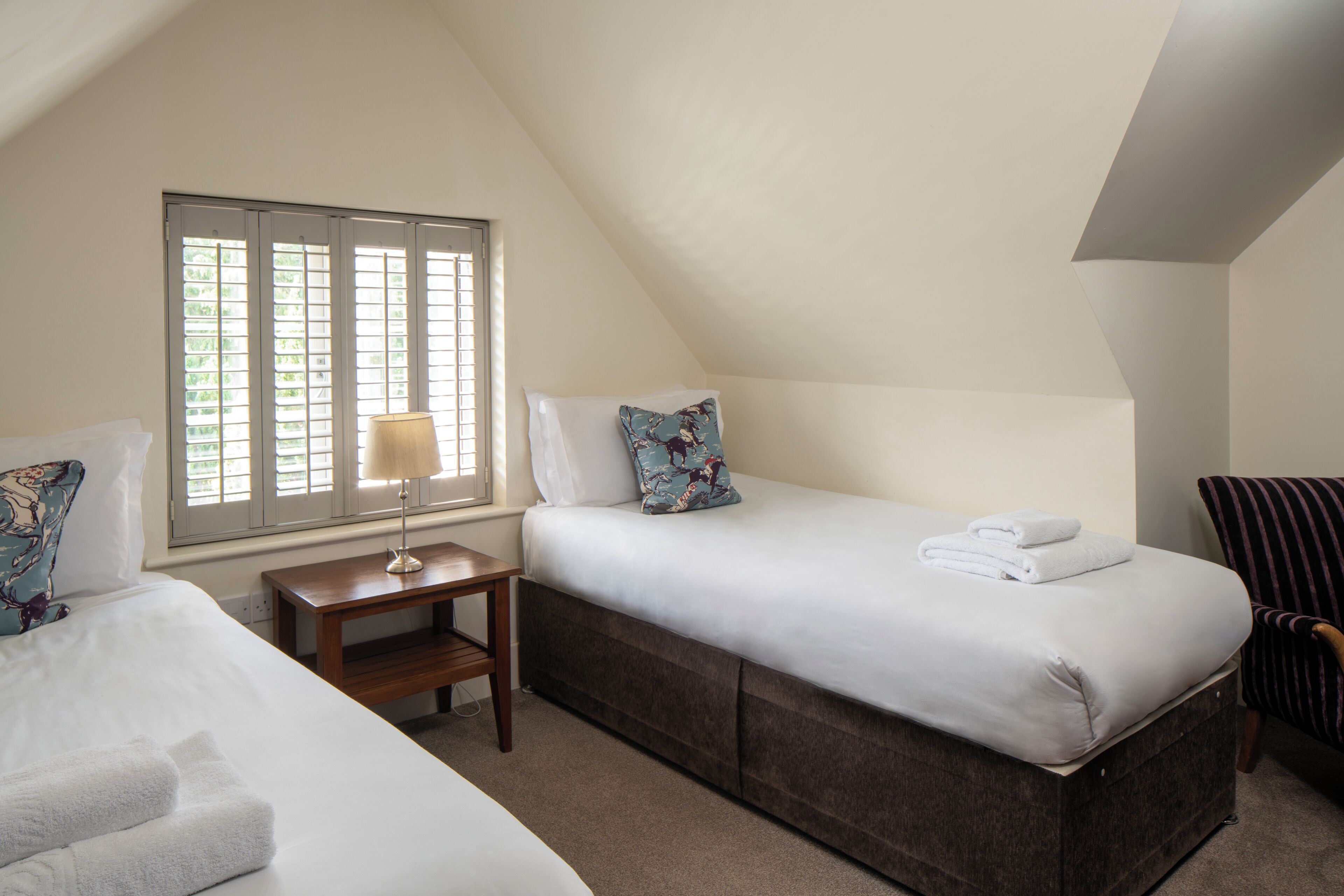 A bedroom with a bed, a bedside table with a lamp, a window with blinds, and a chair.
