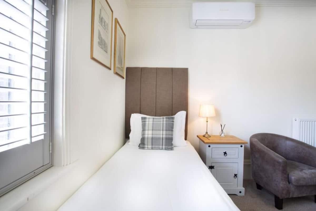 A hotel room with a bed, a nightstand, a lamp, a chair, and a window with blinds.