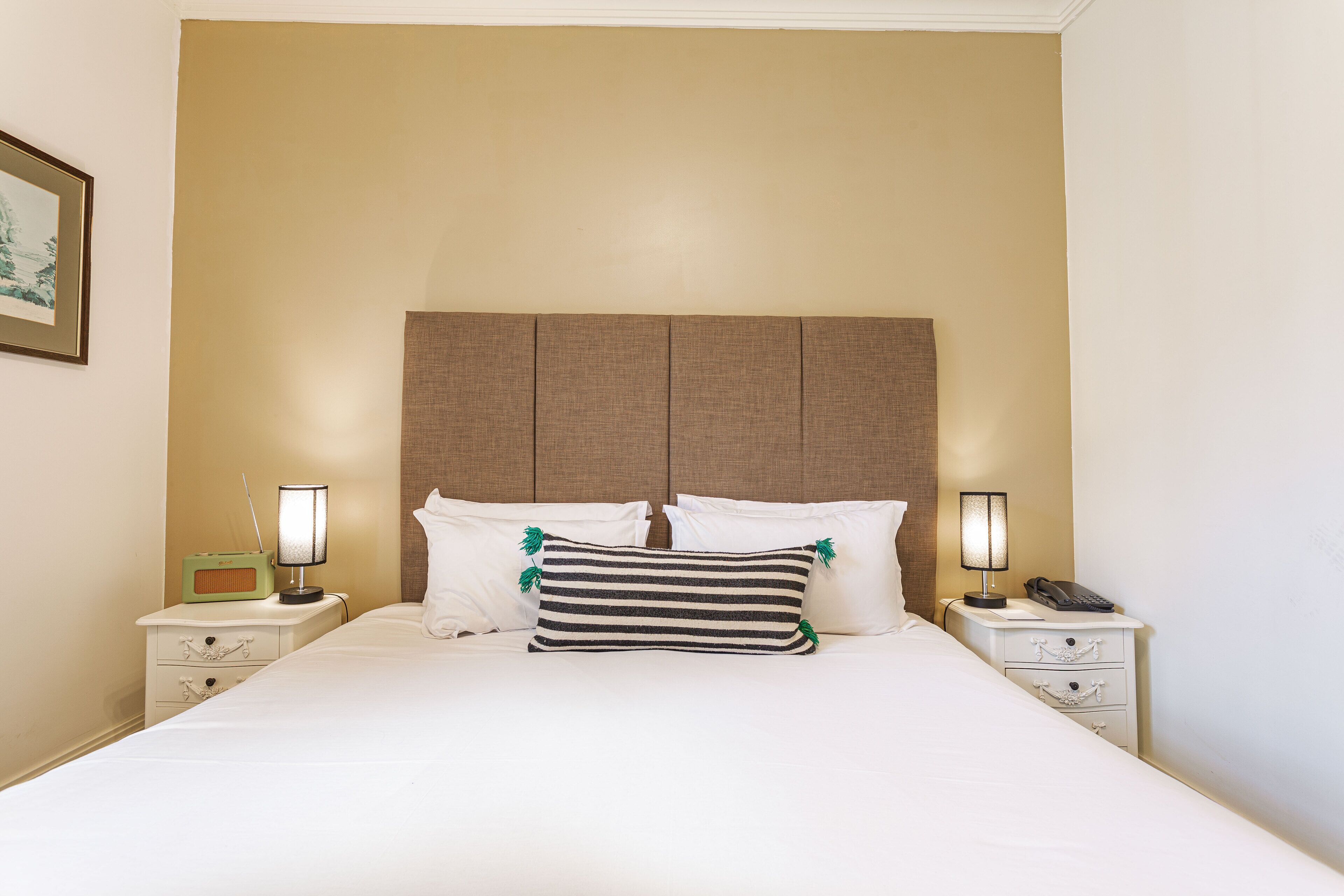 Una cama bien hecha con ropa de cama blanca y almohadas a rayas, flanqueada por dos mesitas de noche con lámparas y un radio.