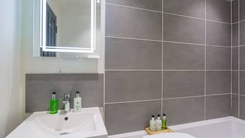 A modern bathroom with a white sink, mirror, and bathtub. There are toiletries on the sink and a wooden tray with a towel on the bathtub.