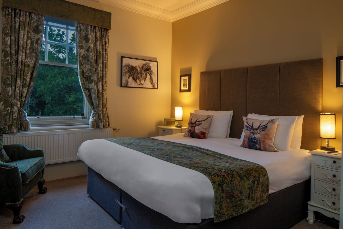 A hotel room with a large bed, a chair, a nightstand with a lamp, and a window with curtains.
