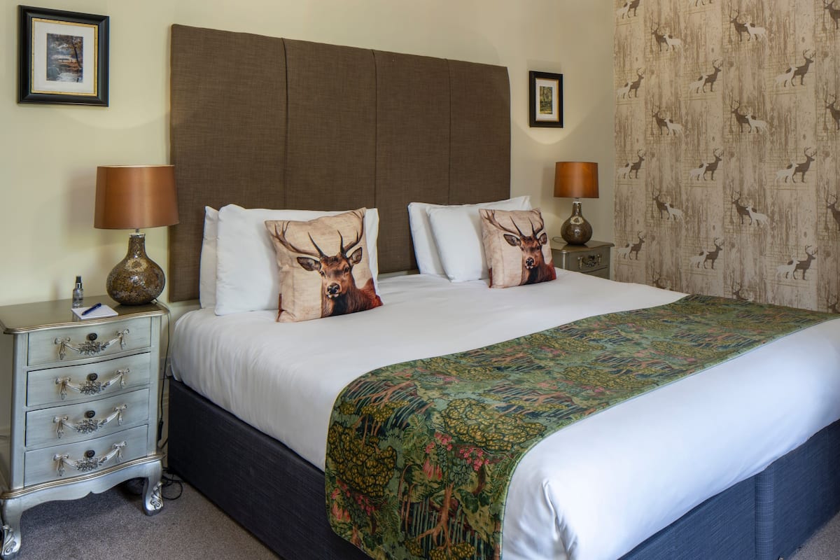 A hotel room with a large bed, two white pillows, a decorative deer pillow, a green floral bedspread, a nightstand with a lamp, and a dresser with drawers.