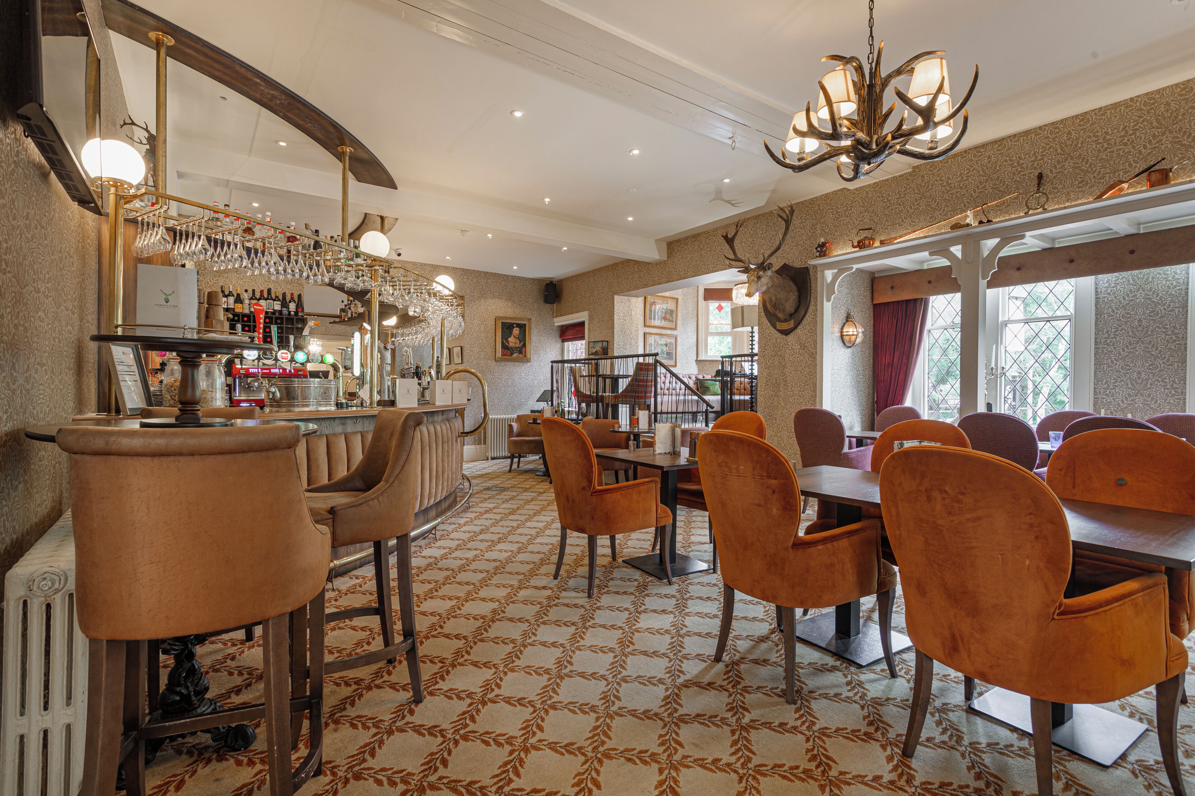 A bar area with a counter, bar stools, and a patterned floor. There are dining tables with chairs, a chandelier, and a large window with curtains.