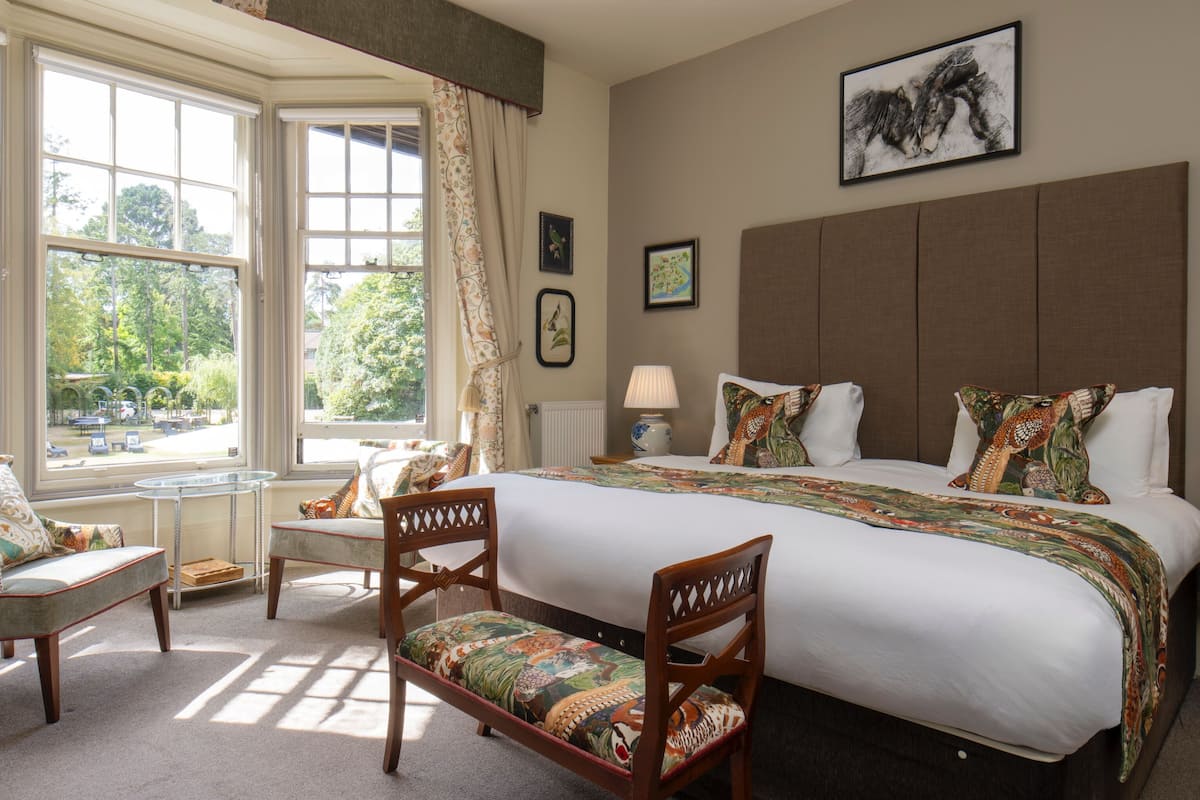 A hotel room with a large bed, two chairs, a bedside table, a lamp, and a window with curtains.