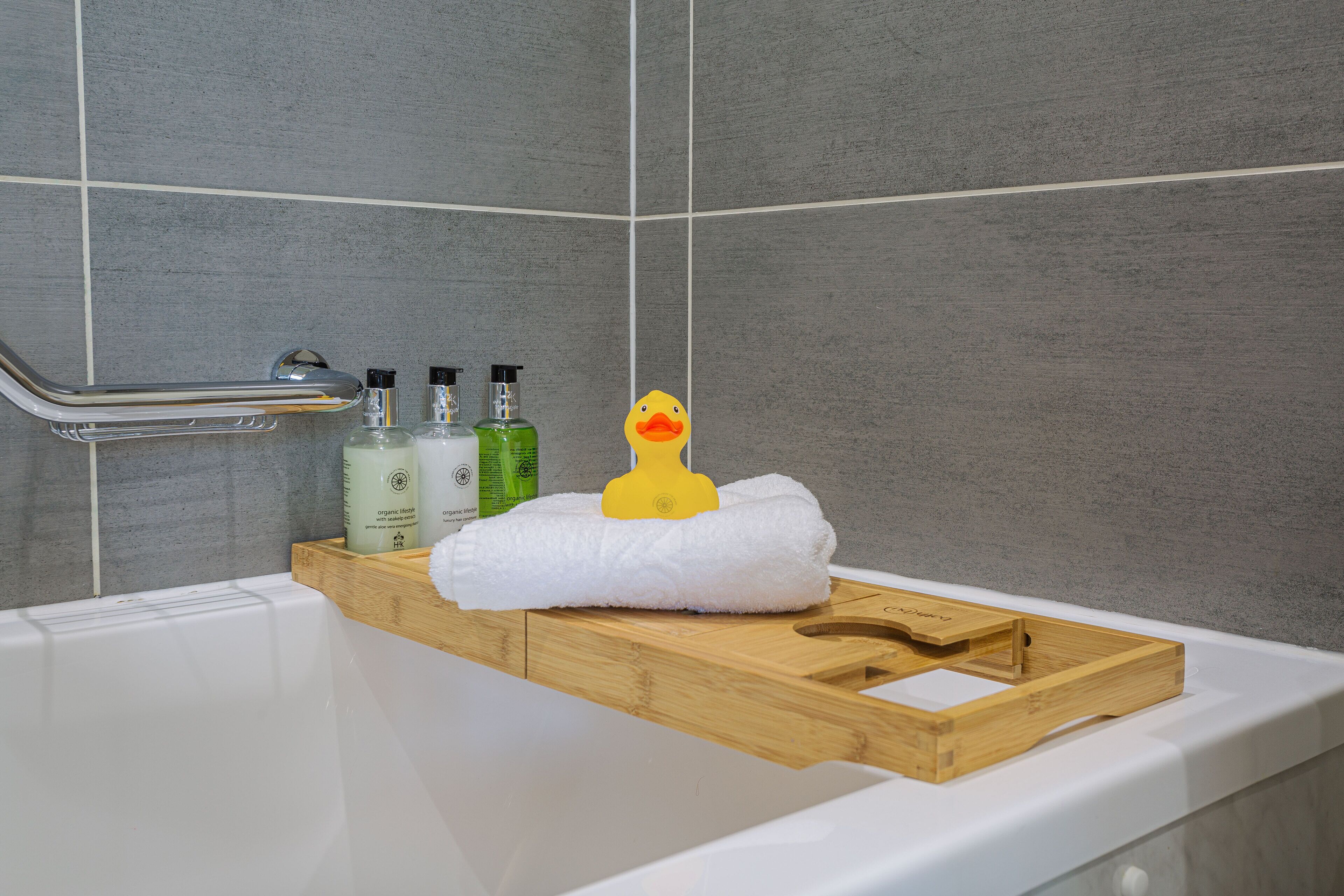 A bathroom with a bathtub, a wooden tray, a white towel, and three bottles of liquid on the side.