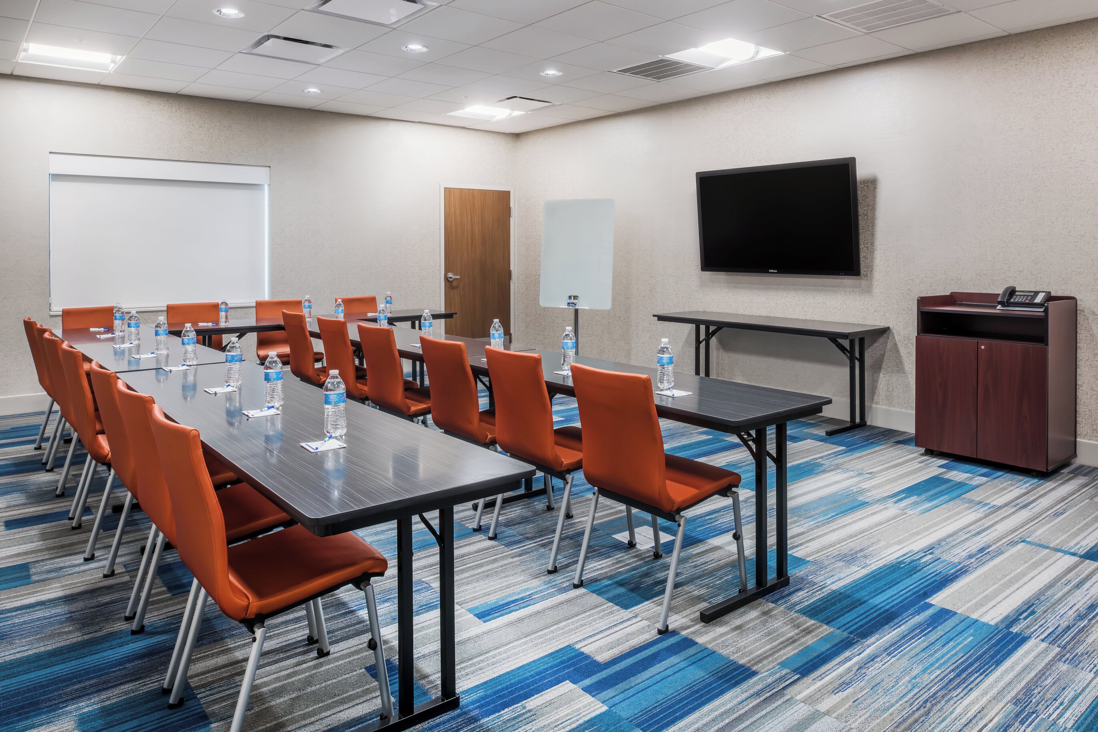 A conference room with a long table, orange chairs, water bottles, a whiteboard, a door, a TV, and a telephone.
