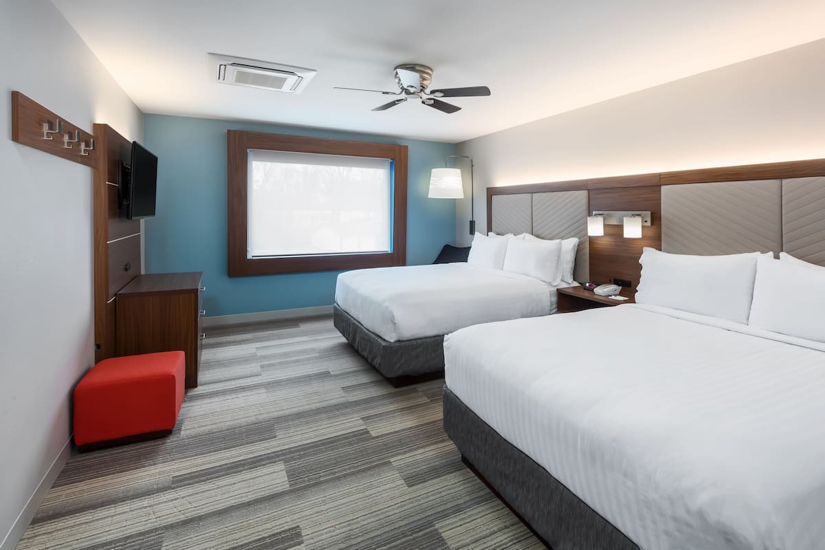 Habitació d'hotel amb dos llits, un pouf vermell, un ventilador de sostre, una televisió i una finestra amb llum natural.