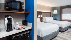 A hotel room with a kitchenette, including a microwave, coffee maker, and tea maker. There are two beds with white bedding and a large window with blinds.