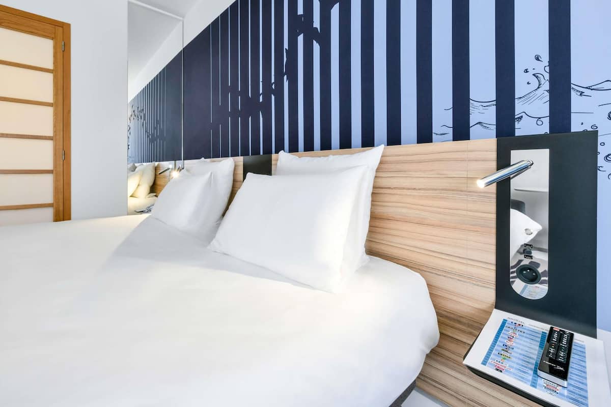 A hotel room with a bed, a wooden headboard, a wall with vertical lines, and a mirror with a remote control.