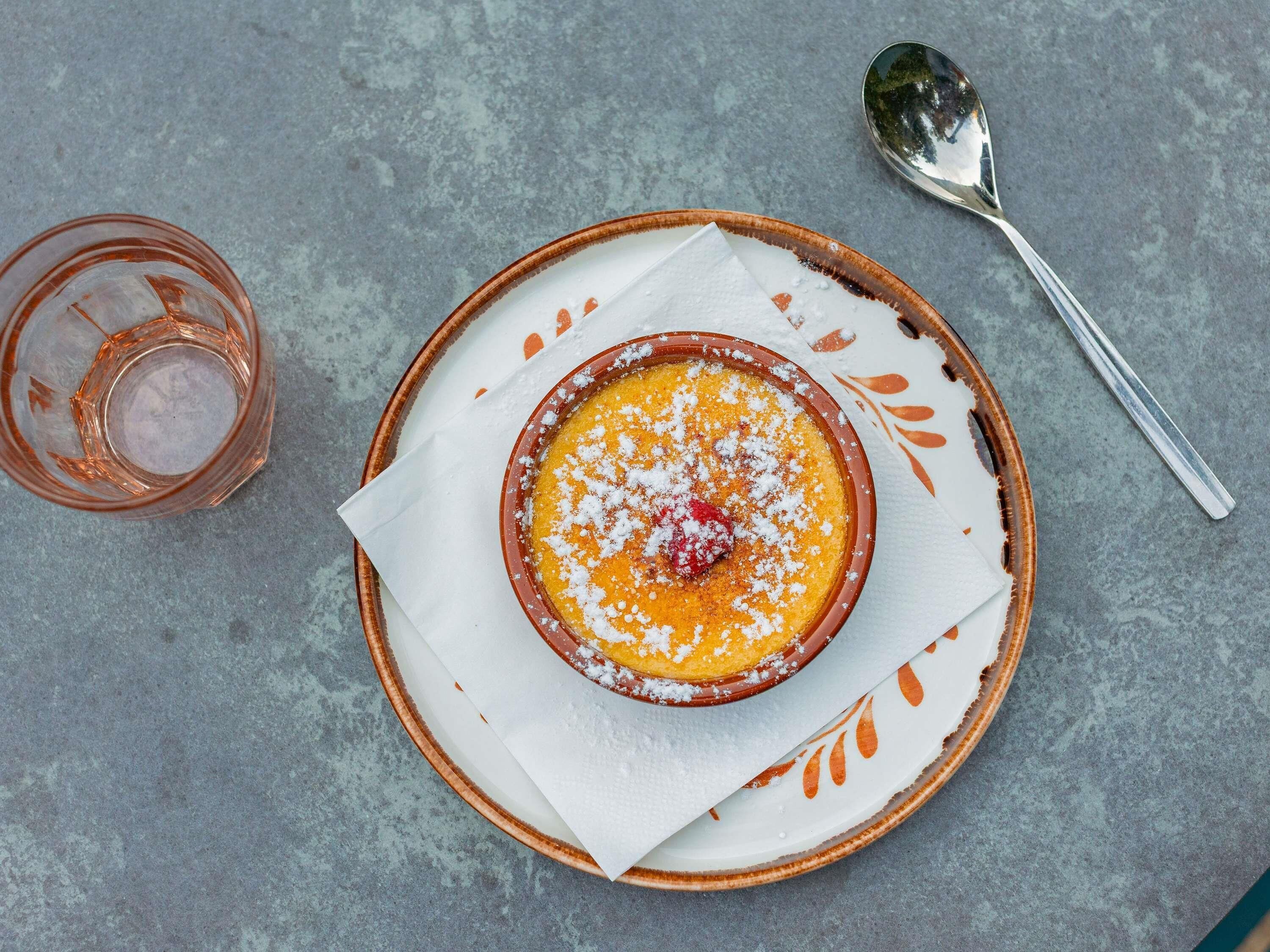 Een dessert in een keramieken schaaltje op een bord, met een lepel en een glas ernaast.