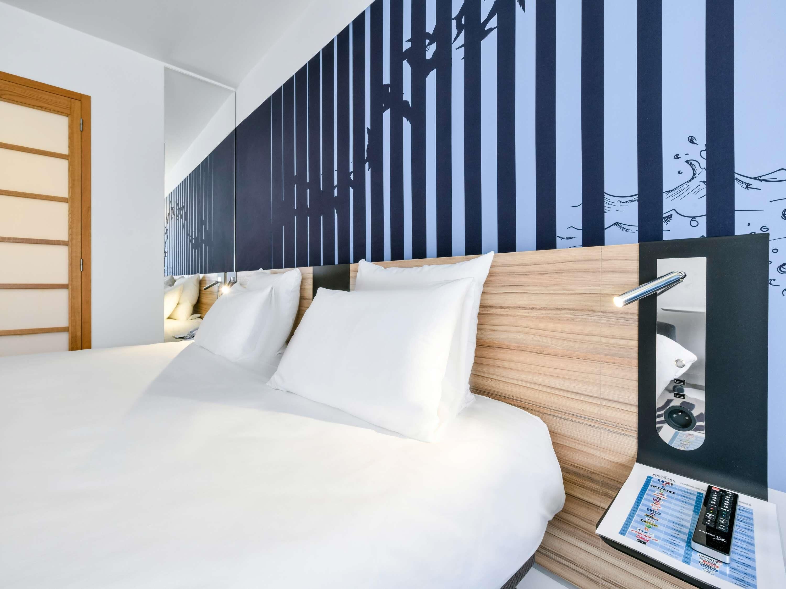 Une chambre d’hôtel avec un lit, une tête de lit en bois, un mur orné de lignes verticales et un miroir doté d’une télécommande.