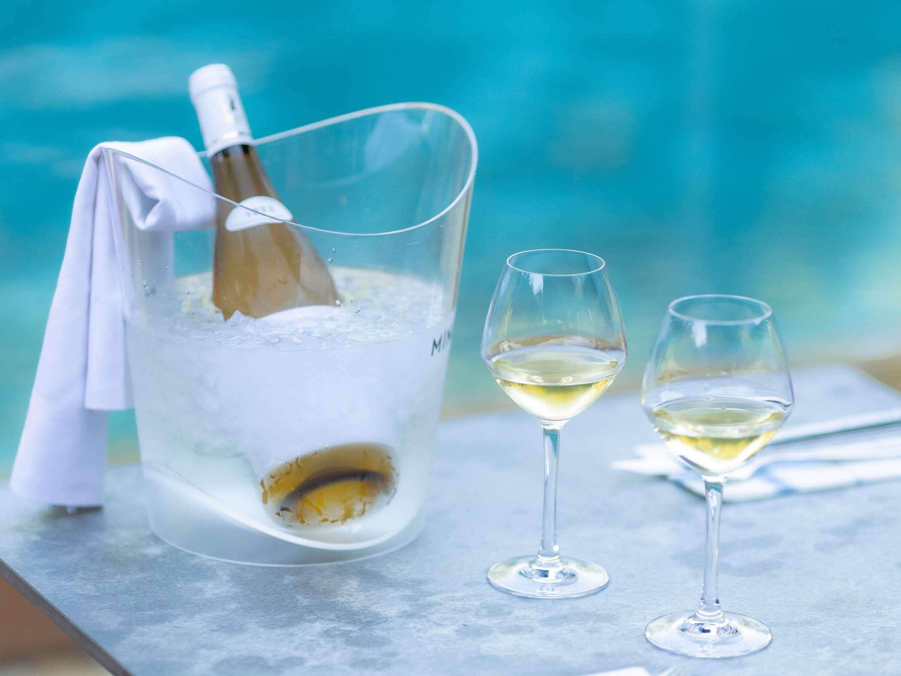 A clear glass bucket with a bottle and ice, two empty wine glasses on a table.