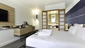 A modern hotel room with a large bed, a flat-screen TV mounted on the wall, a wooden dresser, a built-in desk with a coffee machine, a mirror, and a bedside table with toiletries. - Novotel La Rochelle Centre (La Rochelle)