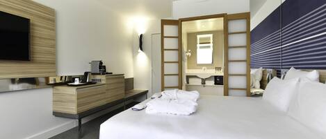 Quarto de hotel moderno com uma cama grande, televisão de tela plana na parede, penteadeira de madeira, escrivaninha embutida com máquina de café, espelho e mesa de cabeceira com produtos de higiene pessoal.