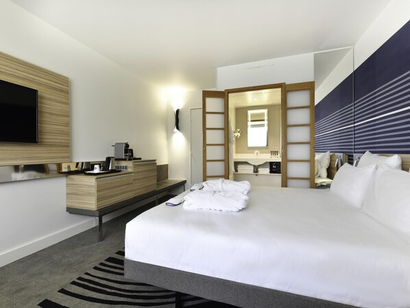 Une chambre d’hôtel moderne dotée d’un grand lit, d’un téléviseur à écran plat fixé au mur, d’une commode en bois, d’un bureau intégré avec une machine à café, d’un miroir et d’une table de chevet avec des articles de toilette.