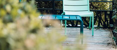 Une chaise sur une terrasse en bois, avec une rambarde en métal et de la verdure au premier plan.